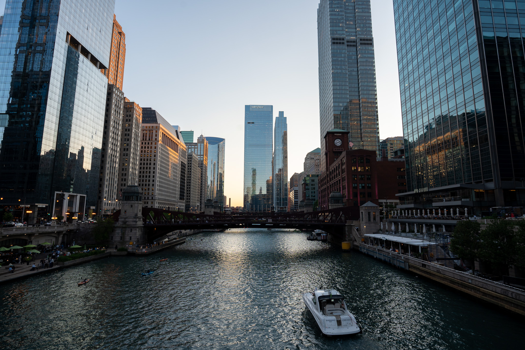 The Chicago Riverwalk at sunset, skyscrapers lining the river with a boat passing beneath a bridge.