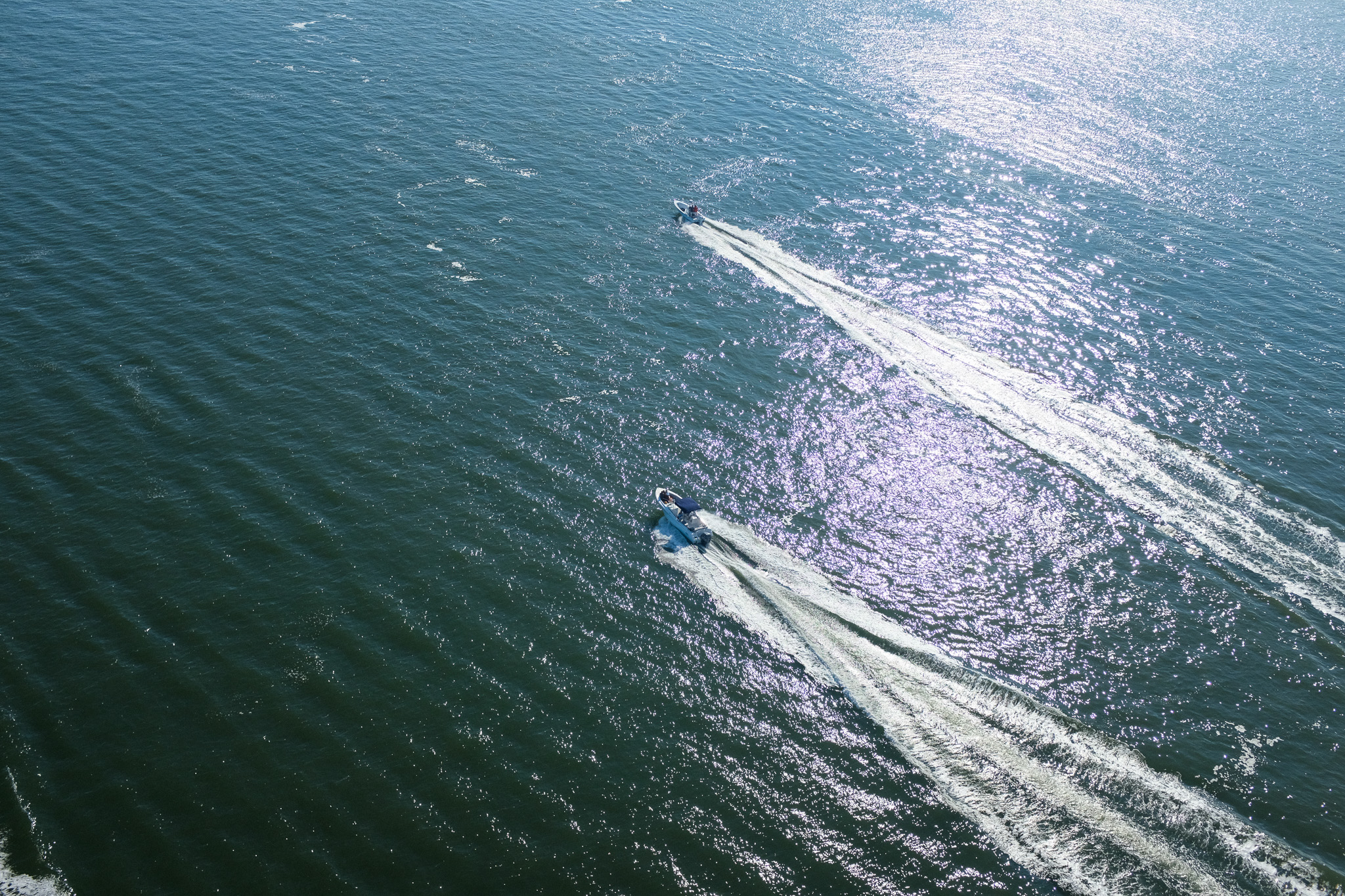 Aerial view of two small motorboats crossing sunlit open blue water, each leaving a long white wake behind it.